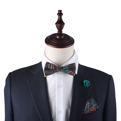 A mannequin in a dark suit and white shirt exudes sophistication with a paisley bow tie, green floral lapel pin, and the Carpe diem Paisley Pocket Square. This elegant ensemble is ideal for weddings, displayed on a crisp white background.