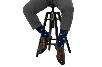 Person wearing gray pants, dark brown dress shoes, and Palm Tree Socks, sitting on a black stool against a plain white background.