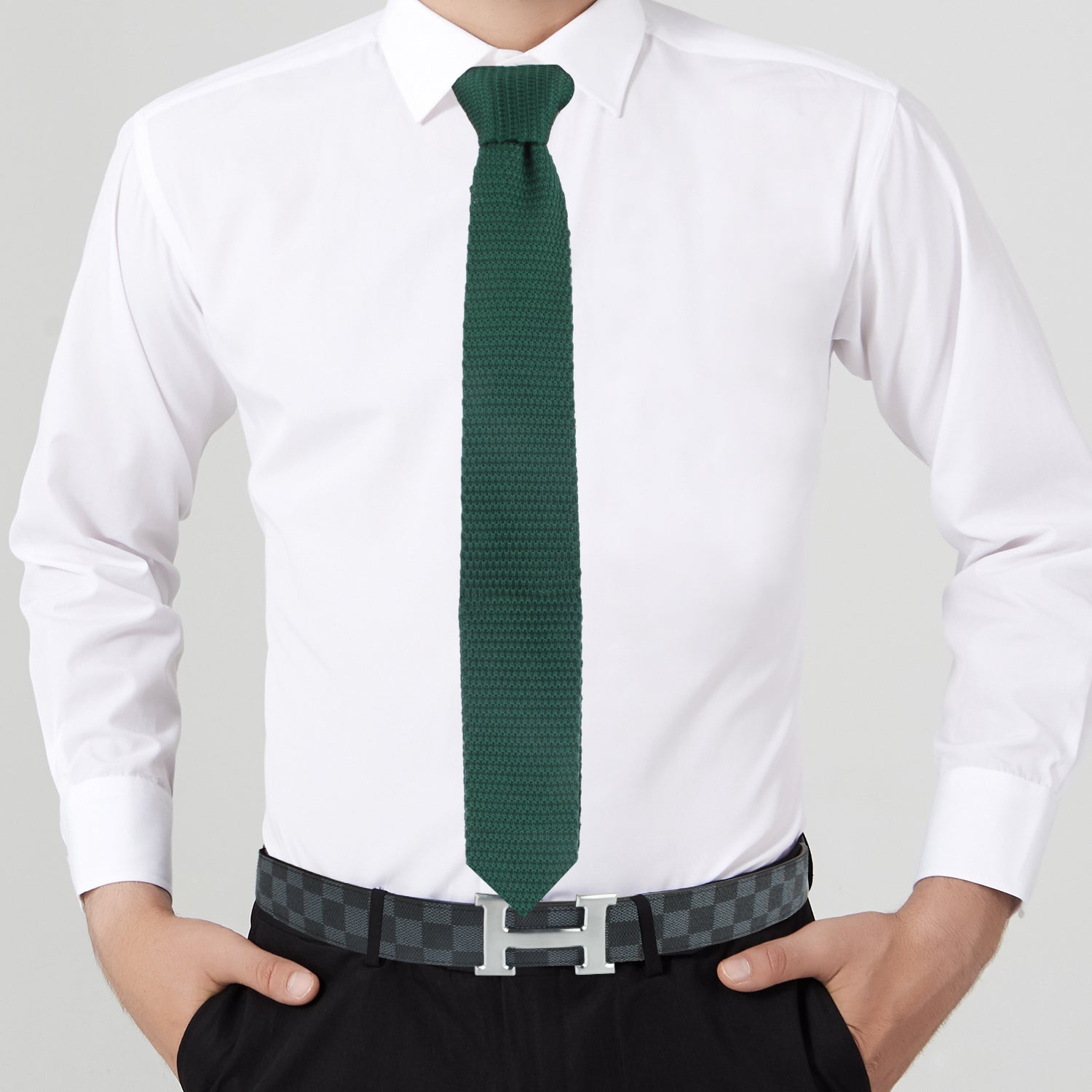 A person exuding elegance in a white dress shirt, Dark Green Knitted Tie, and black pants, accented by a checkered belt featuring a prominent silver "H" buckle.