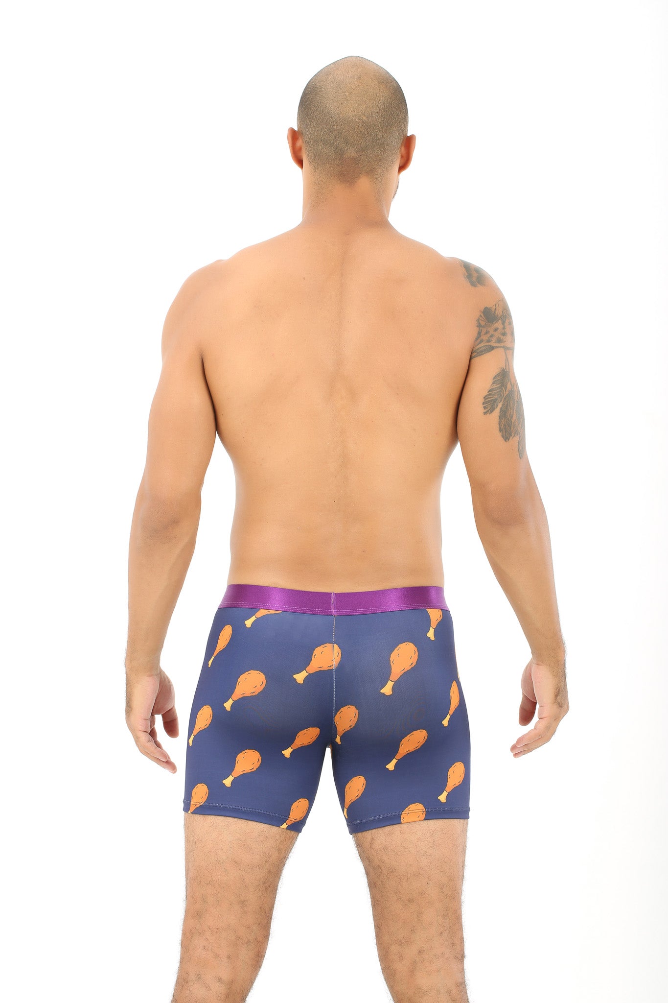 A rear view showcases someone in Drumstick Underwear, featuring quirky orange drumstick patterns against navy blue fabric and a snug purple waistband, oozing cuddly comfort. A tattoo adorns their left arm.