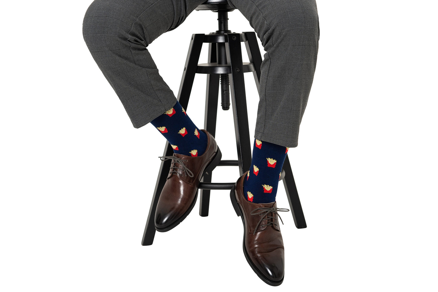 A person in gray plaid pants and brown dress shoes sits on a black stool, wearing Fries Socks featuring a realistic fries print on a navy background.