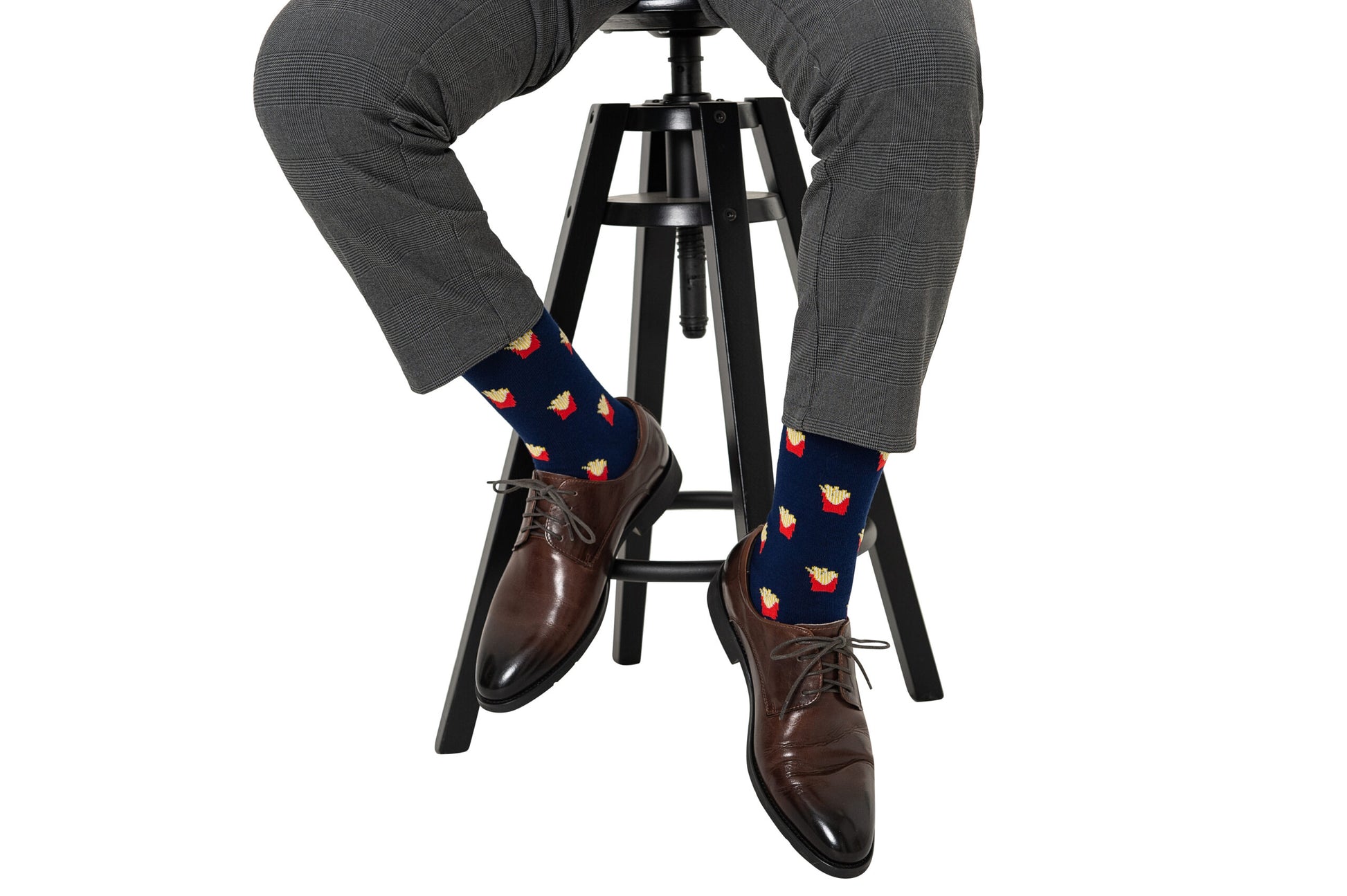 A person in gray plaid pants and brown dress shoes sits on a black stool, wearing Fries Socks featuring a realistic fries print on a navy background.