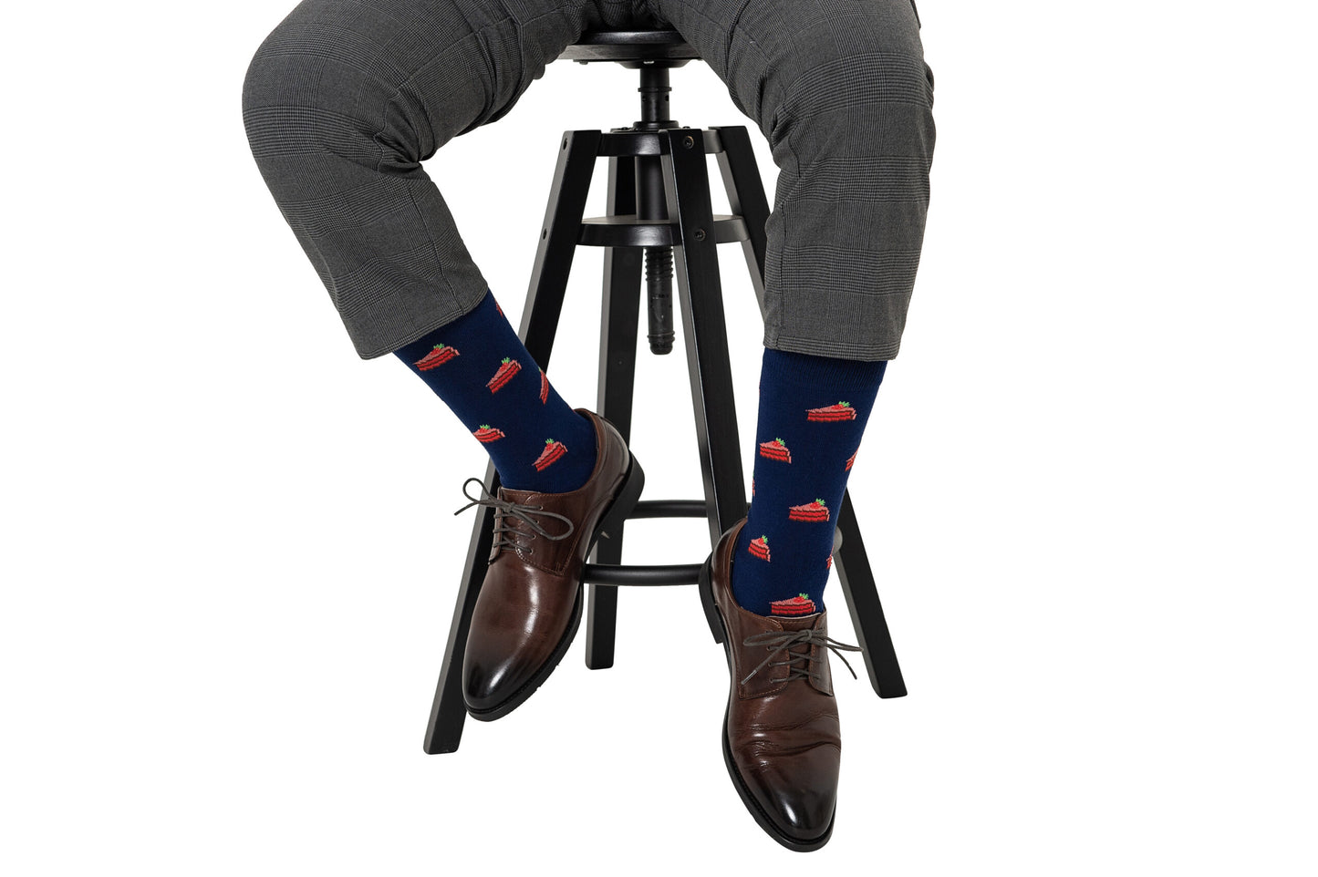 A person wears gray plaid pants, brown dress shoes, and navy blue Strawberry Cake Socks while sitting on a black stool against a white background—Strawberry Cake Socks make a perfect gift for dessert lovers.