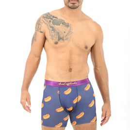 A man stands confidently, showcasing a distinctive style in his Hotdog Boxer—purple boxers decorated with hot dog prints. The tattoo on his left shoulder and arm enhances his appearance, seamlessly combining comfort with personal flair.