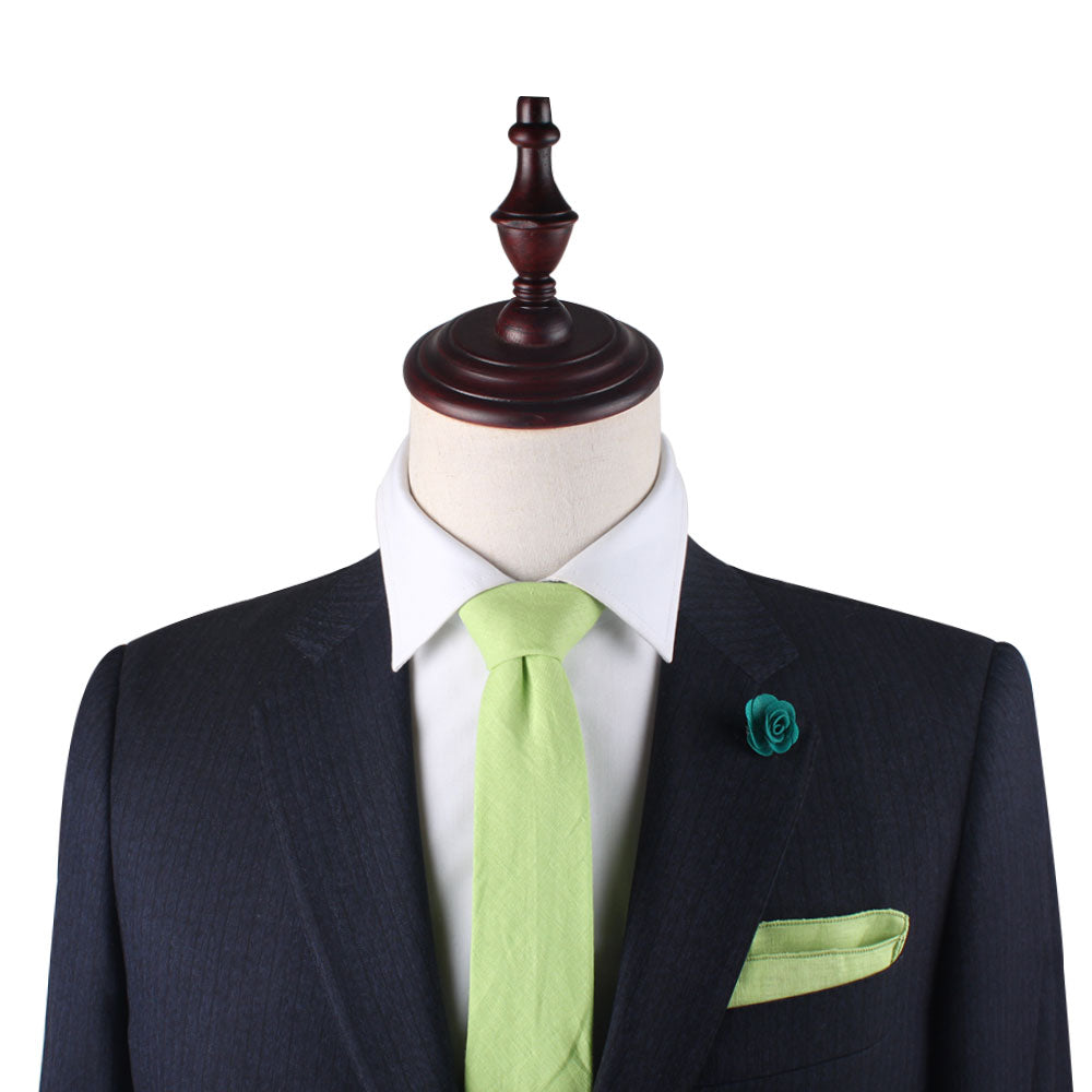 A mannequin dressed in a dark suit with a white shirt, sporting the Lime Green Cotton Skinny Tie & Pocket Square Set, and adorned with a small green flower lapel pin offers a sharp silhouette with zesty lime accents.