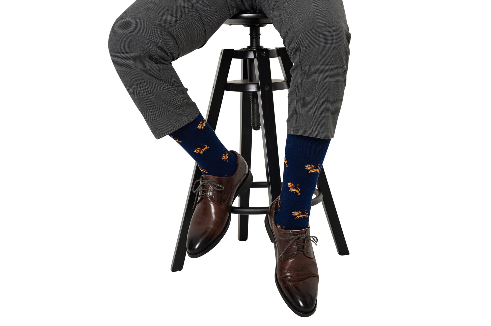 A person wearing gray pants, navy blue Lion Socks with a dog design, and brown leather shoes is seated on a high stool. The style is evident in the meticulous pairing of garments, as the focus zooms in on their legs and feet.