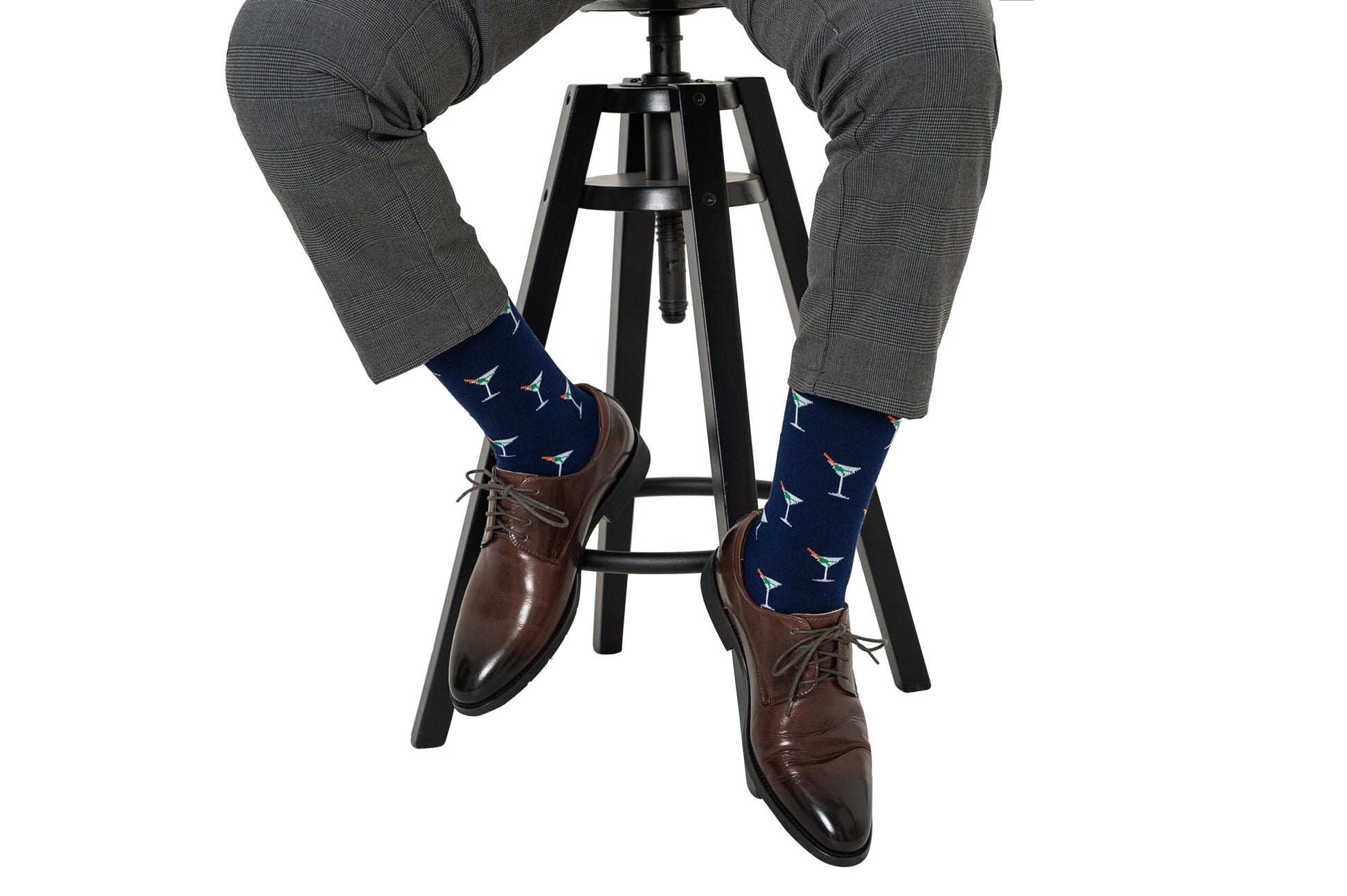 Sporting an impressive sock game, the person sits on a stool in gray pants and brown shoes, showcasing vibrant Martini Margarita Socks adorned with playful cocktail glass motifs.