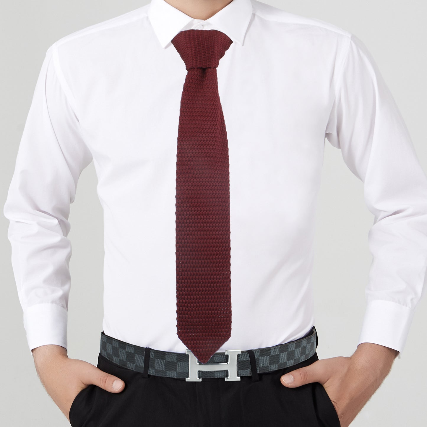 A person wearing a white dress shirt paired with a Mens Knitted Tie and a patterned belt. With their hands in their pockets, the warm-hued background enhances the cozy elegance of their ensemble.