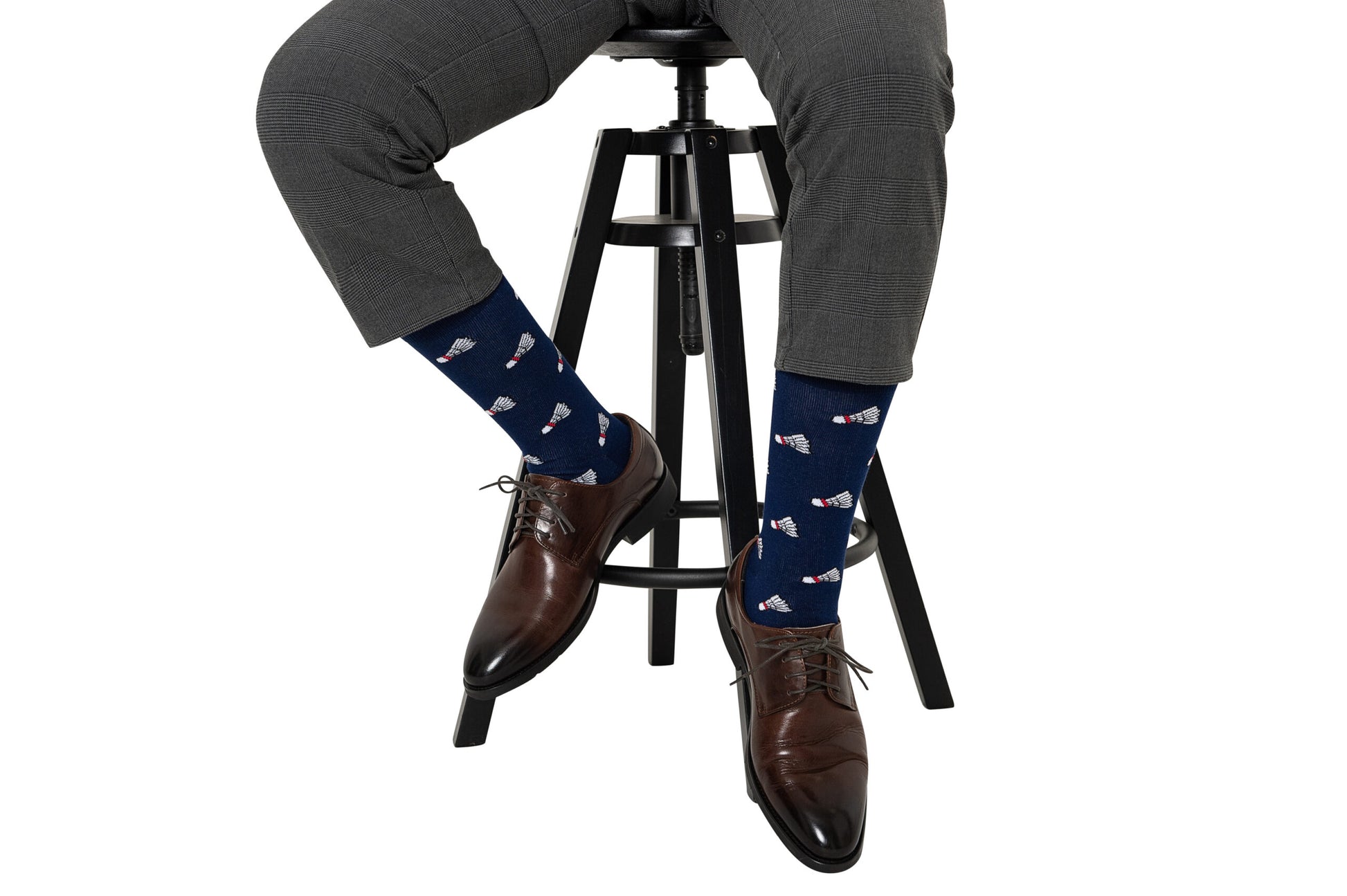 Wearing moisture-wicking Badminton Socks in navy with a red, white, and blue pattern, a person in gray dress pants and brown shoes sits on a stool.