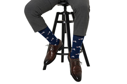 Wearing moisture-wicking Badminton Socks in navy with a red, white, and blue pattern, a person in gray dress pants and brown shoes sits on a stool.