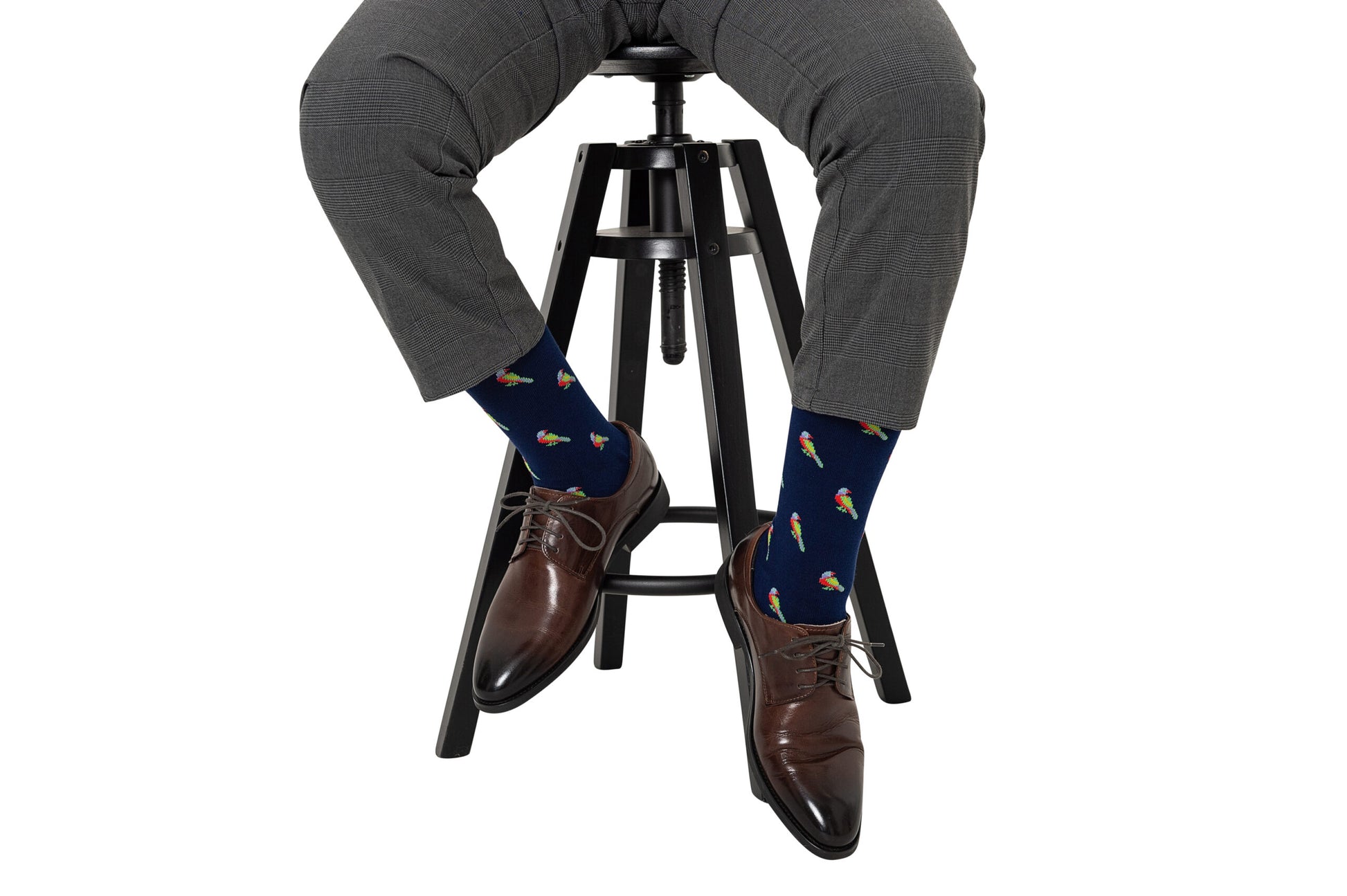 A person in gray pants is seated on a high stool, wearing brown dress shoes and Parrot Socks, which feature colorful tropical parrot designs.