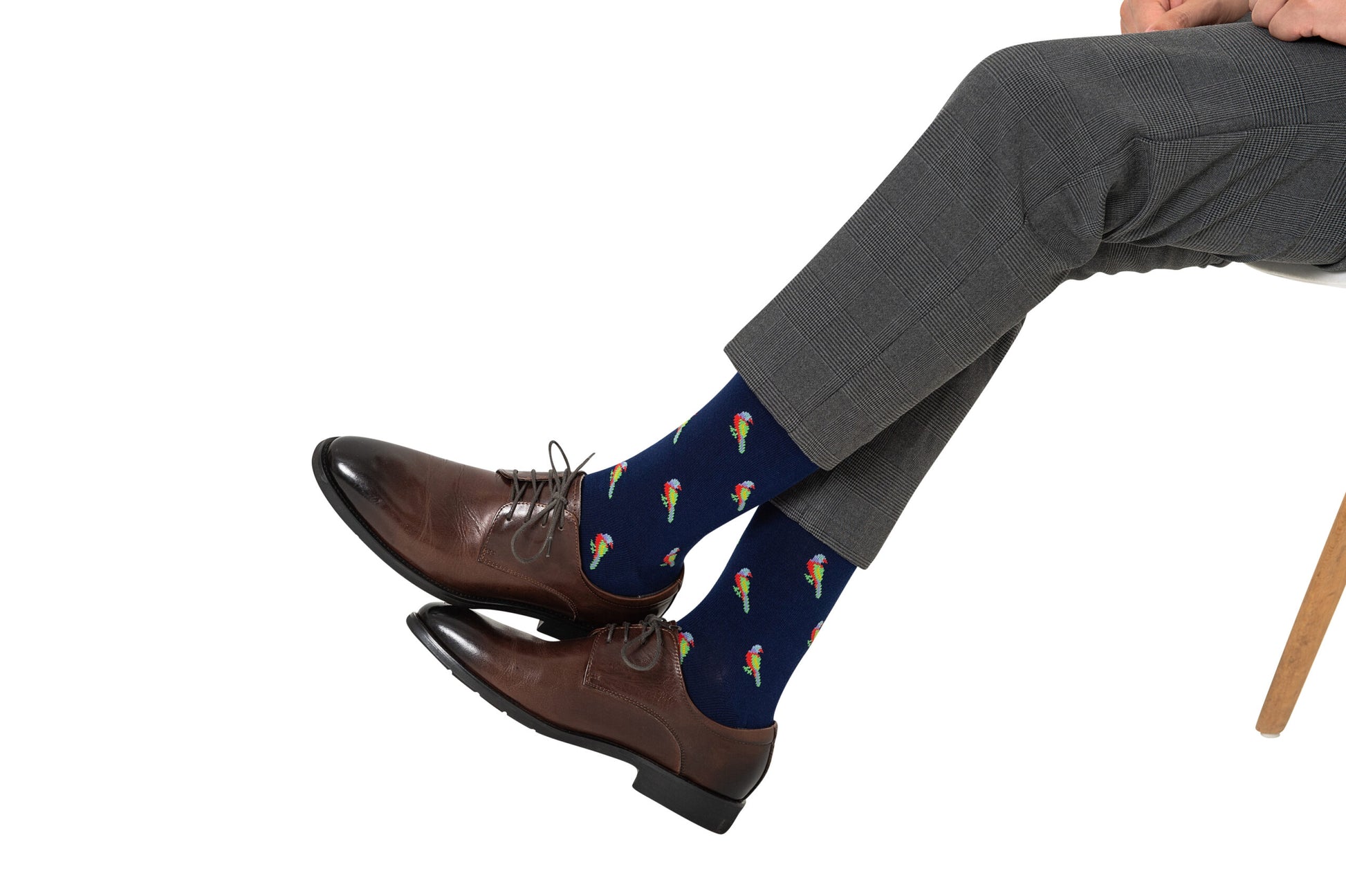 A person seated with legs crossed is wearing gray pants and brown shoes, showcasing vibrant Parrot Socks.