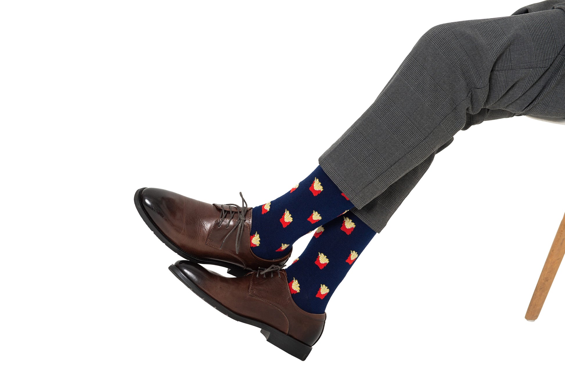 Wearing gray dress pants and brown shoes, a person sits with crossed legs, showing off Fries Socks featuring a realistic fries print.