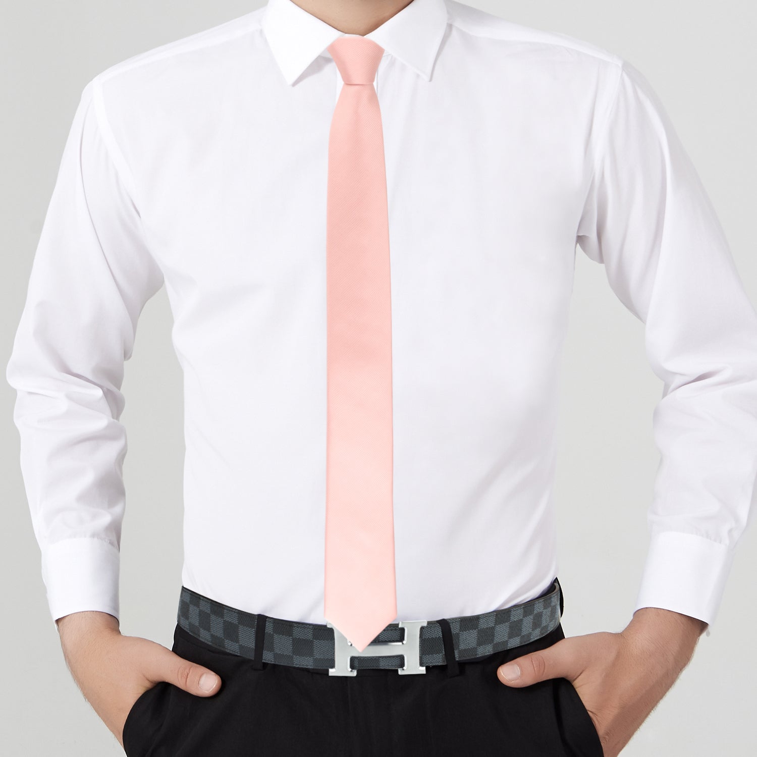Person exuding modern elegance in a white dress shirt, sleek rose gold skinny necktie paired with a matching pocket square, black pants with a checkered belt, standing confidently with hands in pockets.
