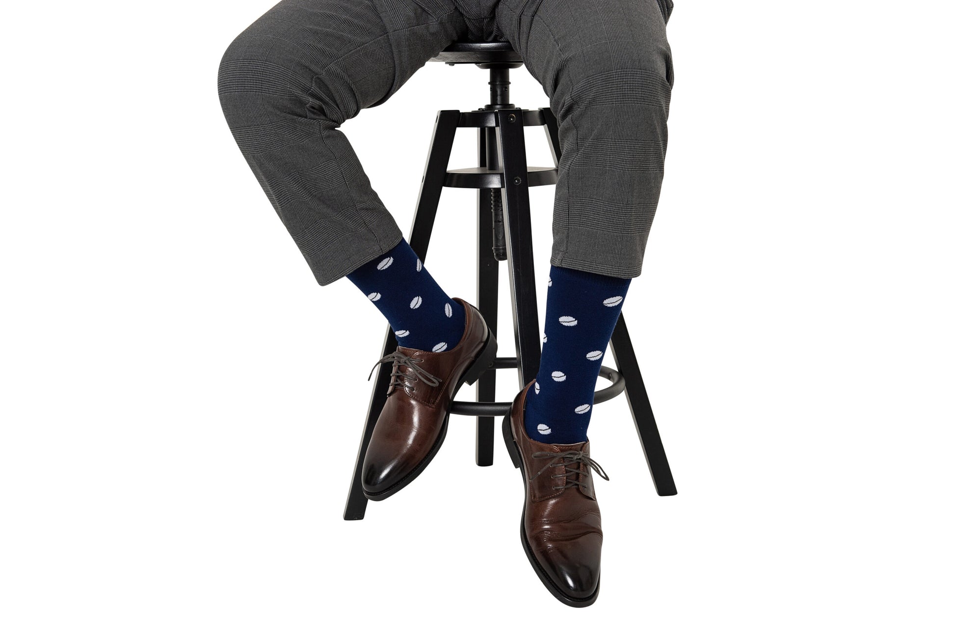 A person seated on a black stool, wearing grey trousers and brown leather shoes paired with Rugby Socks adorned with white polka dots, ready to tackle the day.