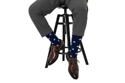 A person seated on a black stool, wearing grey trousers and brown leather shoes paired with Rugby Socks adorned with white polka dots, ready to tackle the day.