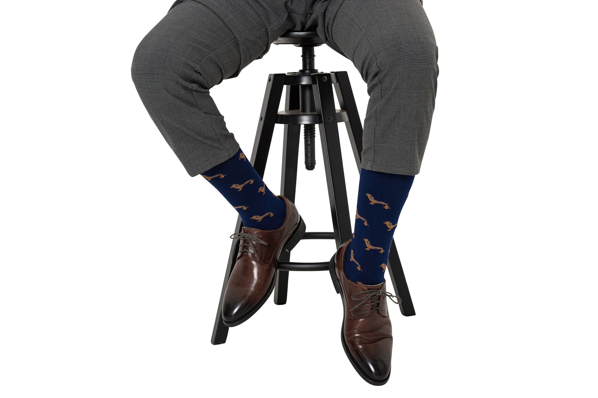 A person wearing gray pants, brown dress shoes, and quirky Sausage Dog Socks sits on a black stool.