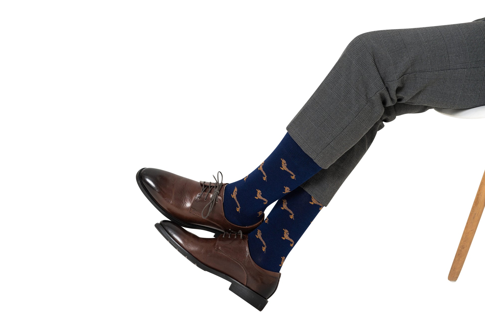 A person wearing brown dress shoes, grey pants, and Sausage Dog Socks featuring quirky brown dog patterns sits with legs crossed on a white background.