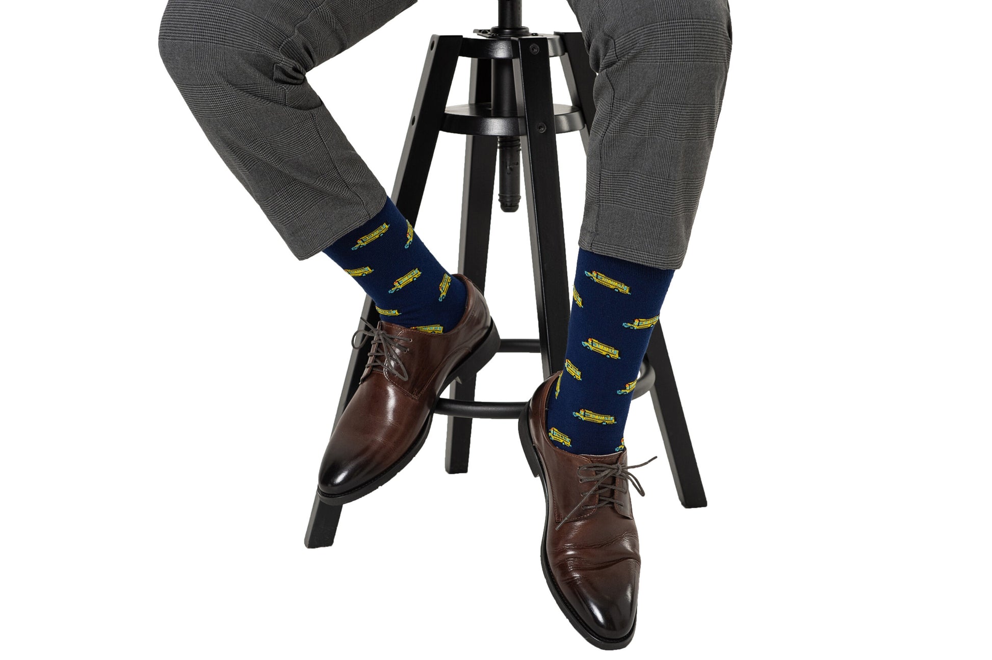 Person wearing gray dress pants and brown leather shoes, sitting on a black stool, showing off their School Bus Socks with blue and yellow airplane patterns that evoke a sense of nostalgia.