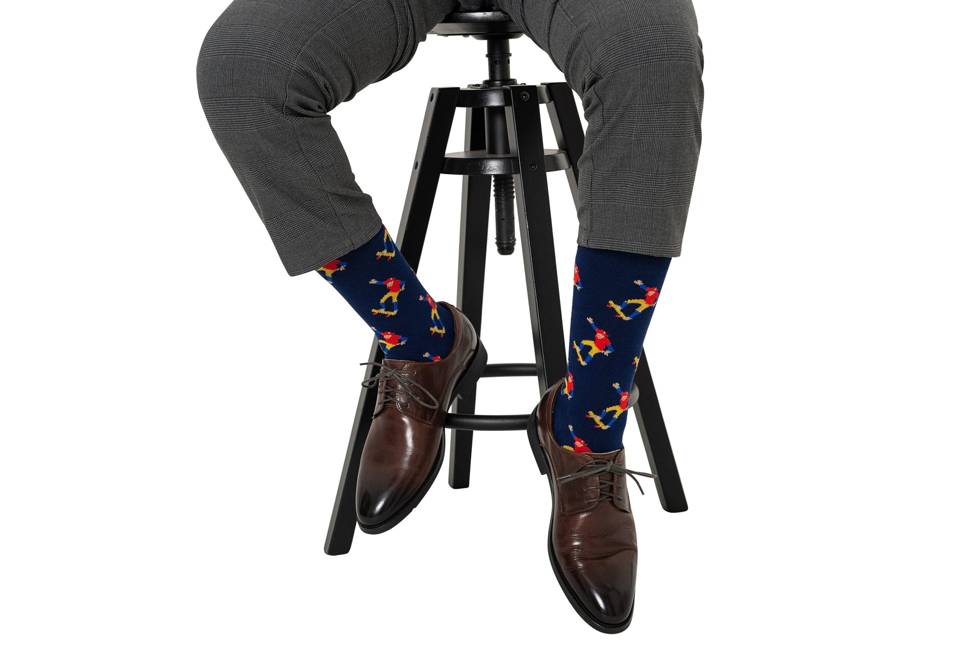 A street-smart individual, seated on a stool, wearing grey checkered pants, brown dress shoes, and Skateboarder Socks featuring a colorful bird pattern.