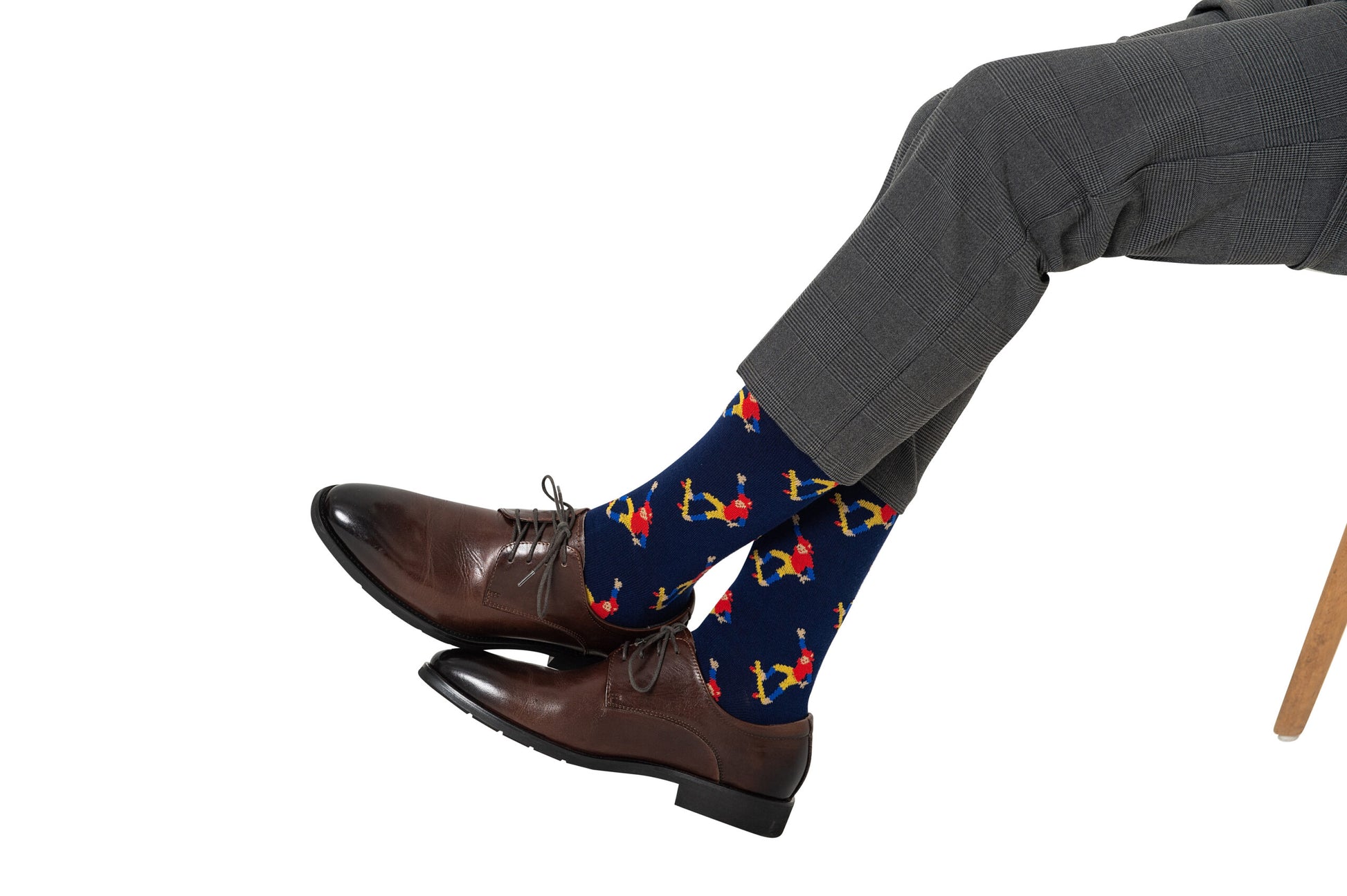 A person exuding street-smart comfort is wearing gray dress pants and Skateboarder Socks with a bartender motif, paired with brown dress shoes, sitting with one leg crossed over the other.