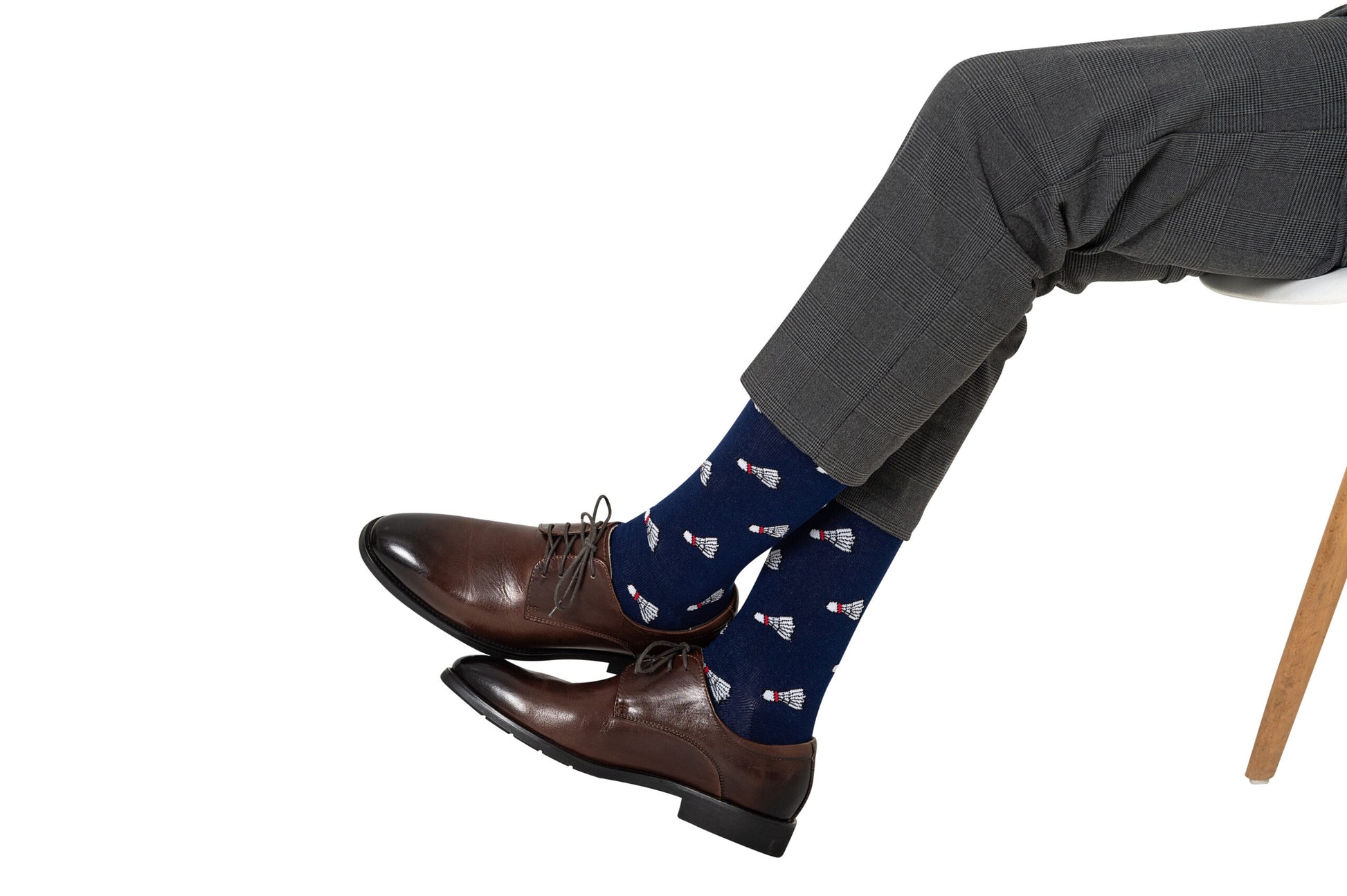 A person wearing Badminton Socks featuring a shuttlecock print is seated on a chair, dressed in gray dress pants and brown dress shoes.