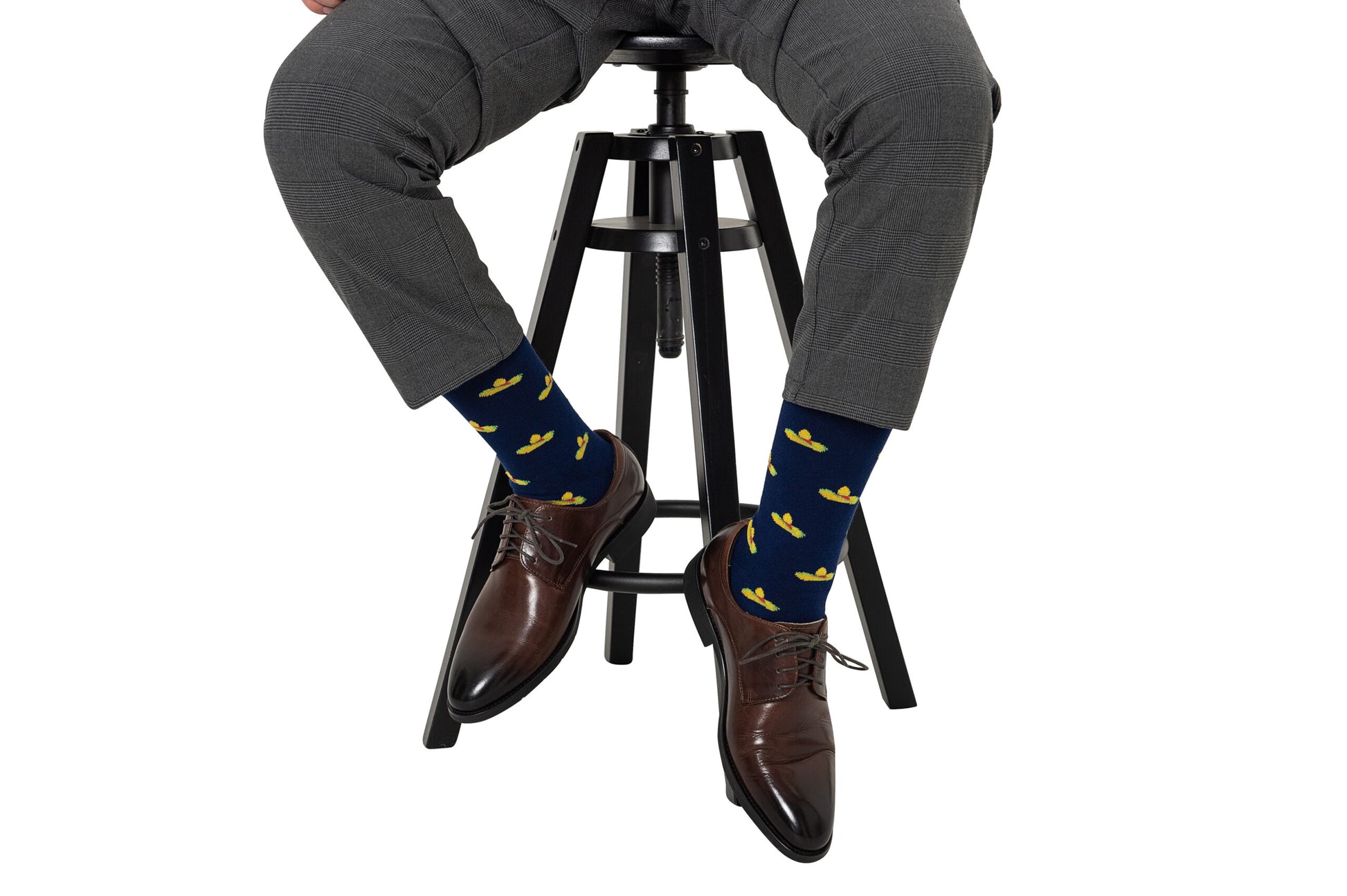 A person in gray pants sits on a black stool, their feet adorned with brown shoes and Sombrero Socks featuring a playful fiesta of yellow duck patterns.