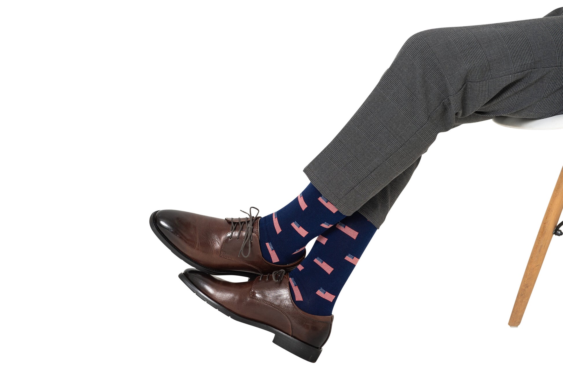 A man in gray trousers is sitting, wearing brown leather shoes and USA Flag Socks that add a touch of style.
