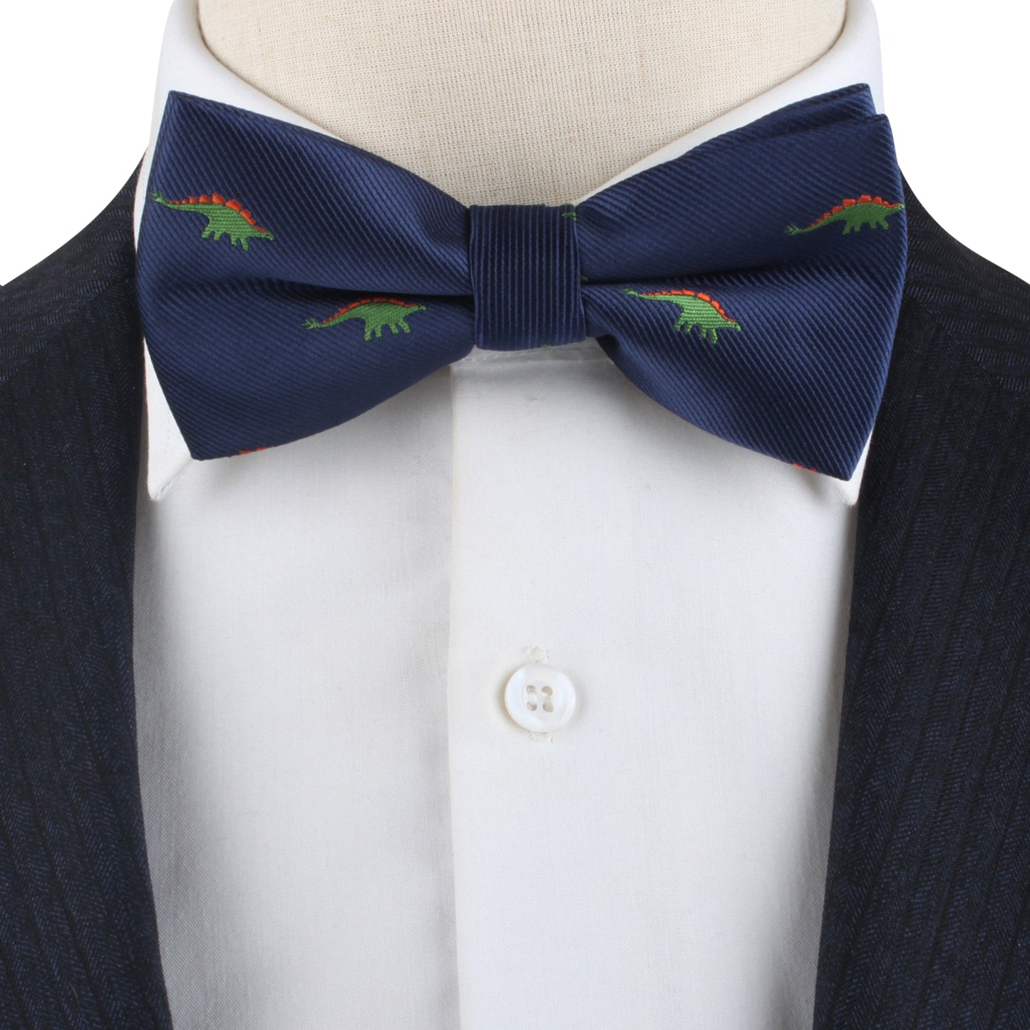 A close-up of the Stegosaurus Bow Tie, featuring a navy blue fabric adorned with small green and red dinosaur patterns, worn with a white button-up shirt and a dark jacket. This accessory adds a touch of prehistoric panache to the ensemble.