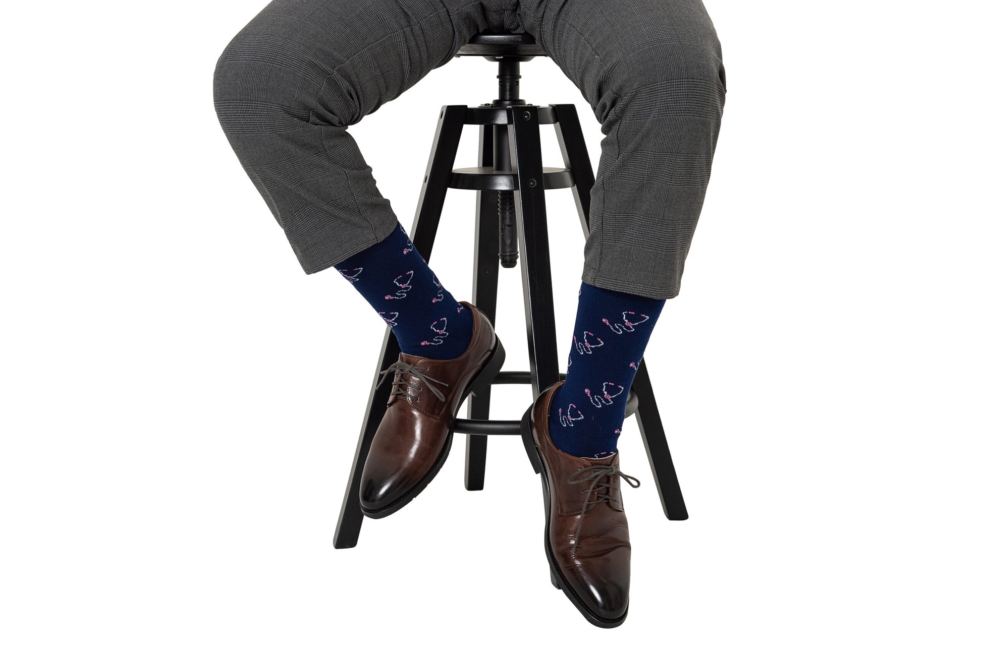 A person wearing gray pants, brown dress shoes, and Stethoscope Socks featuring heart patterns sits on a black stool against a white background.