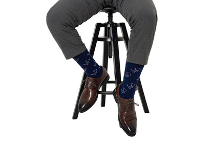 A person wearing gray pants, brown dress shoes, and Stethoscope Socks featuring heart patterns sits on a black stool against a white background.