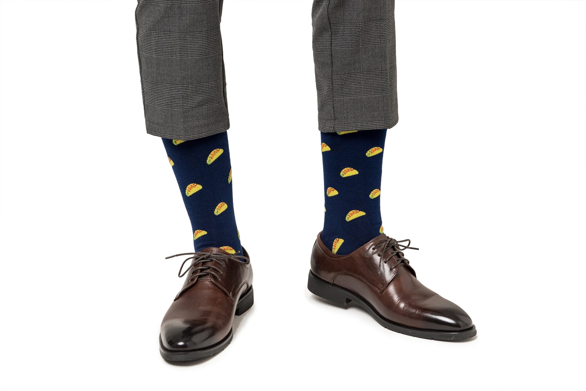 A person wearing gray trousers, blue Taco Socks with yellow taco patterns that add a flavor-packed playfulness, and brown leather dress shoes.