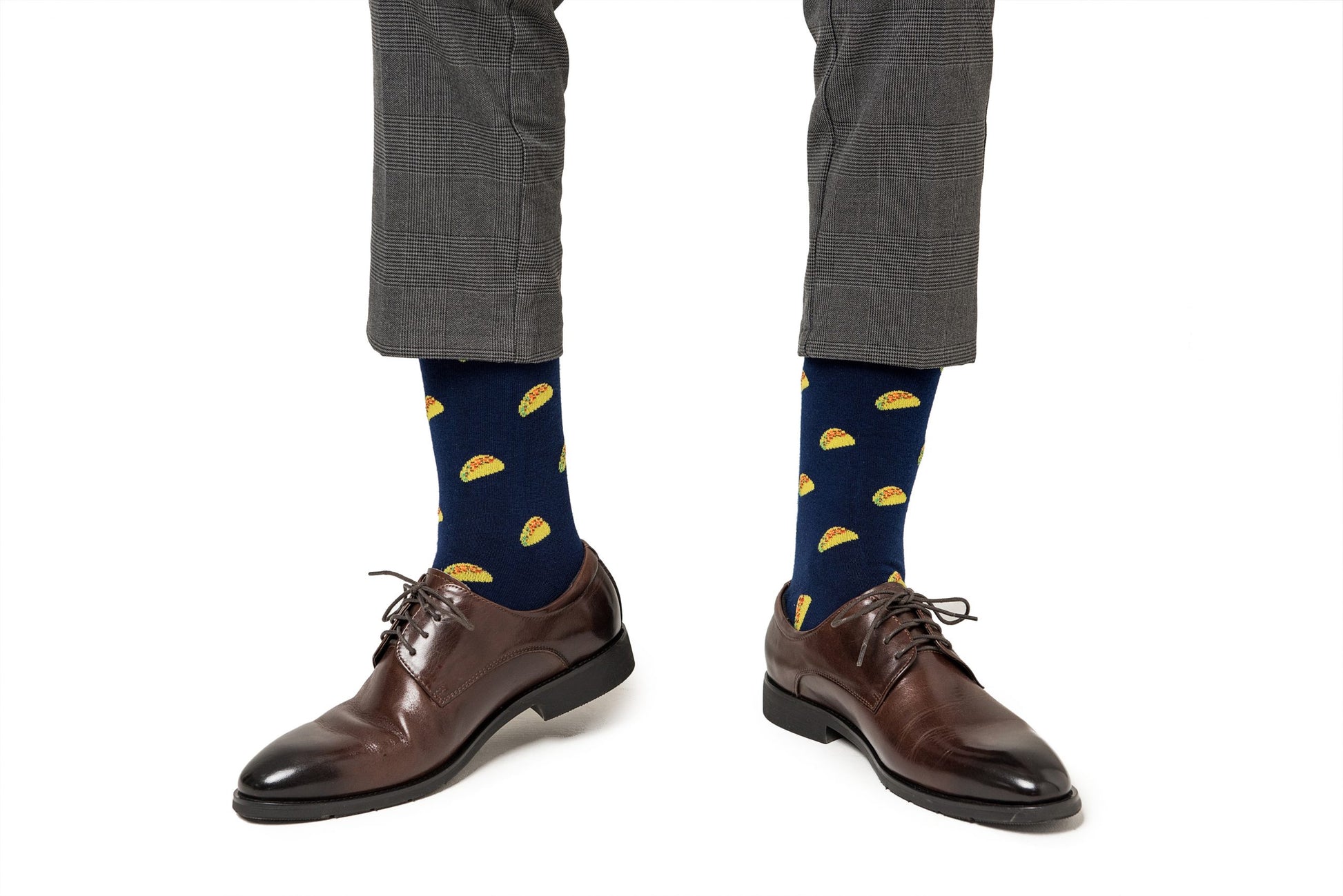 A person wearing dark brown leather shoes and Taco Socks adorned with flavor-packed yellow taco prints, paired with grey trousers.