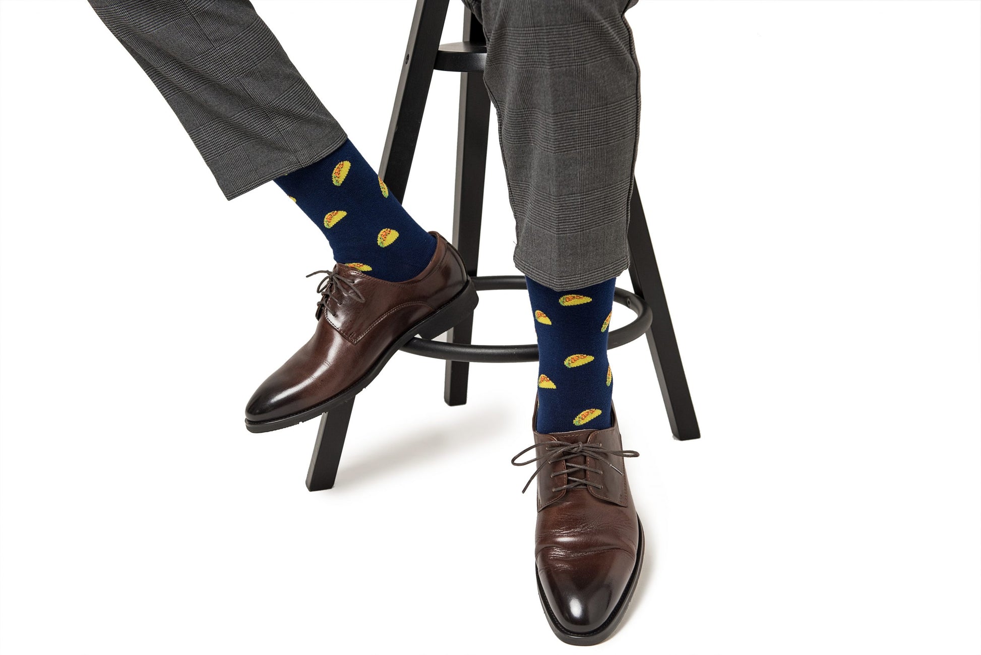 A person sits on a stool wearing brown leather shoes and flavor-packed Taco Socks with lemon patterns, paired with grey trousers.