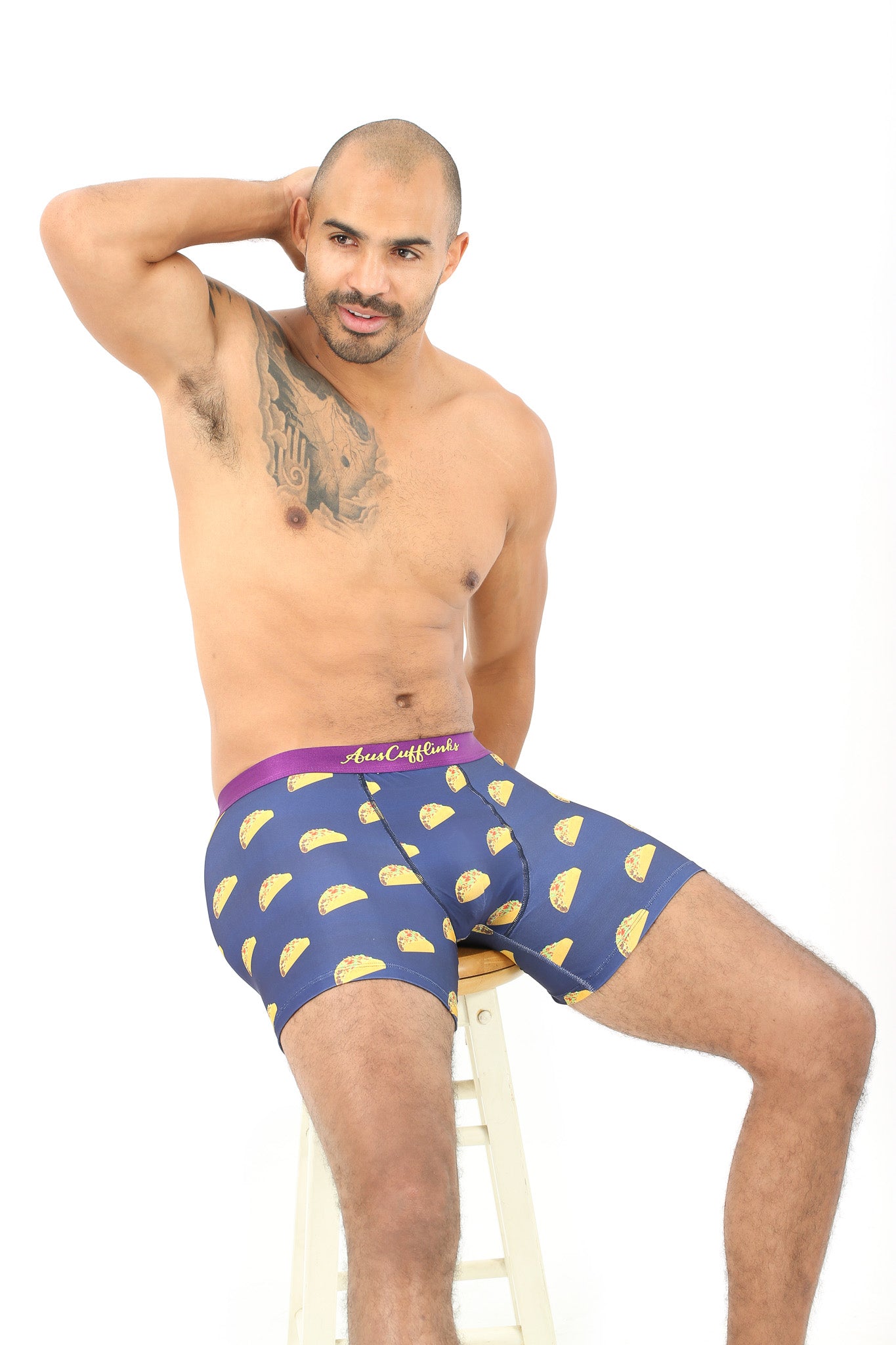 A tattooed man sits on a stool wearing Taco Underwear—blue boxers with taco patterns. Perfect for taco enthusiasts, these moisture-wicking boxers offer all-day comfort.