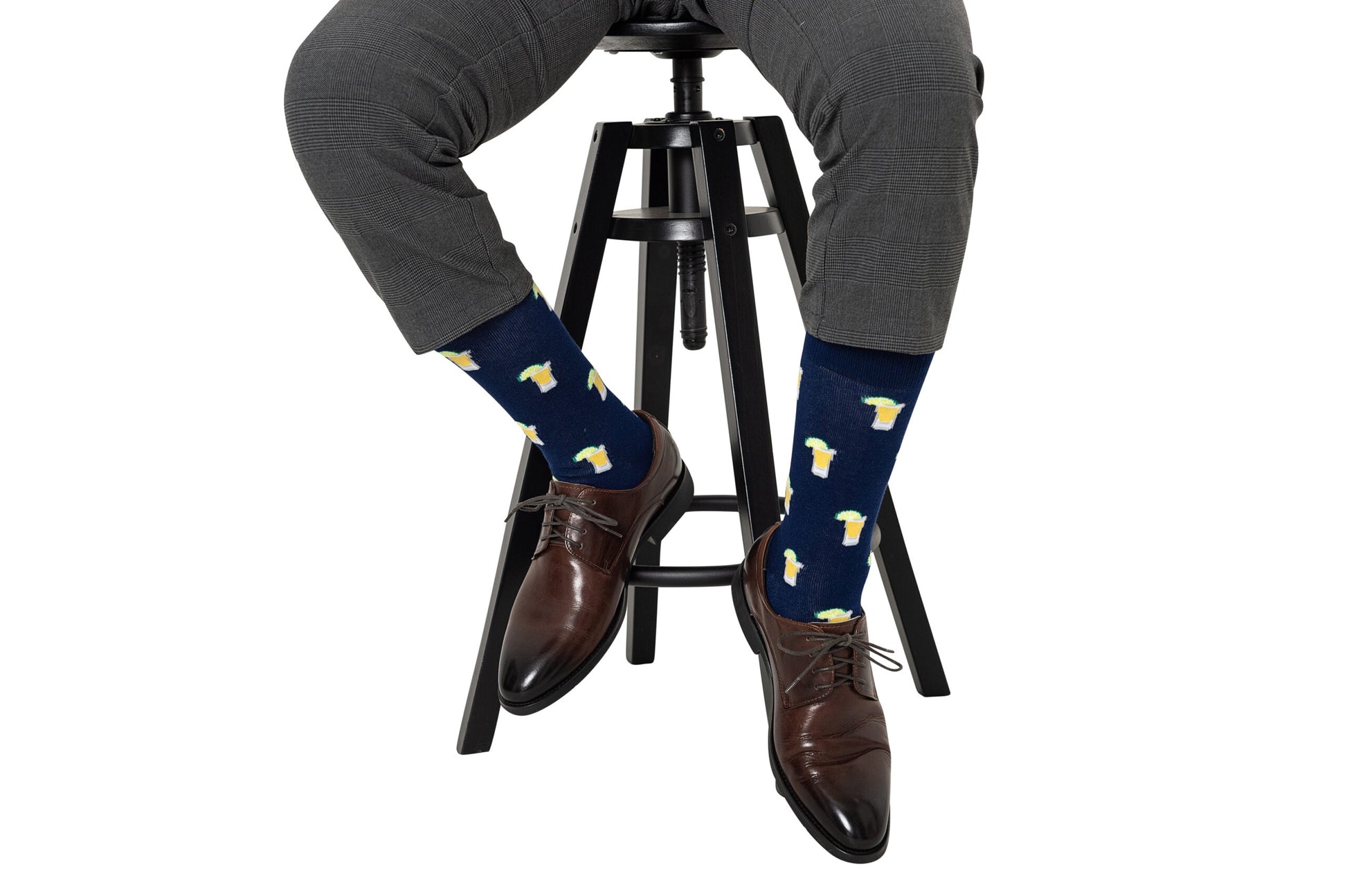 A person seated on a stool exudes a spirited style, wearing gray trousers, Tequila Socks with yellow and white accents, and brown dress shoes.