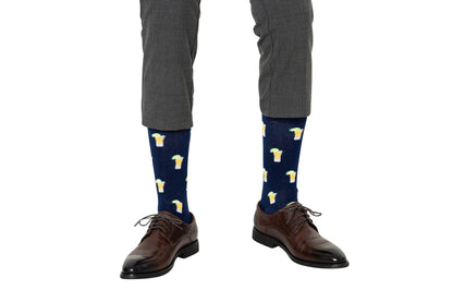 A person wearing gray dress pants, dark brown dress shoes, and Tequila Socks—navy blue socks with beer mug patterns—stands against a white background, perfect for tequila lovers who want fun party socks.