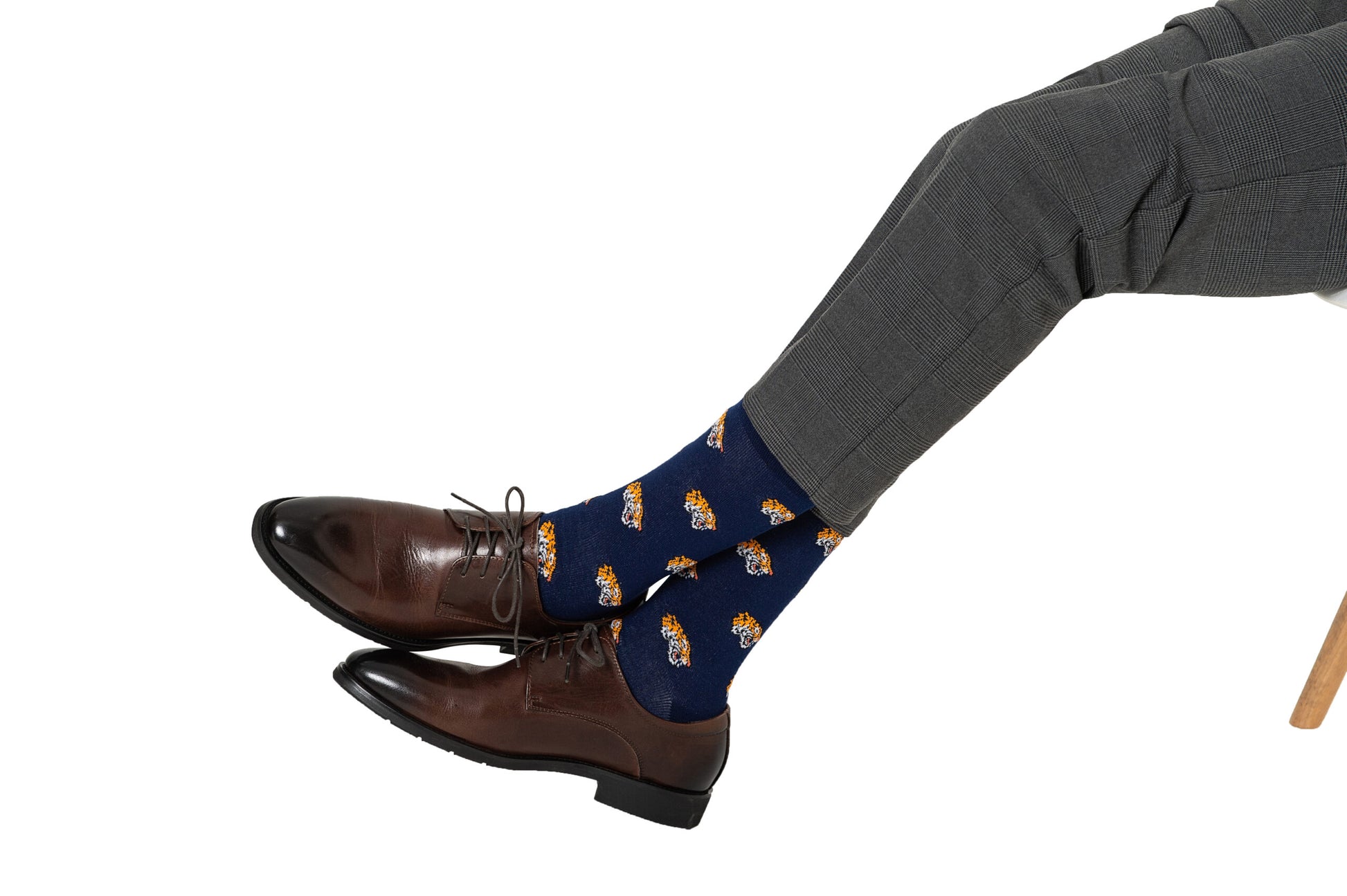 A person in gray pants is sitting with legs crossed, wearing brown dress shoes and blue socks with a colorful pattern that brings a touch of Tiger Socks style to their ensemble.