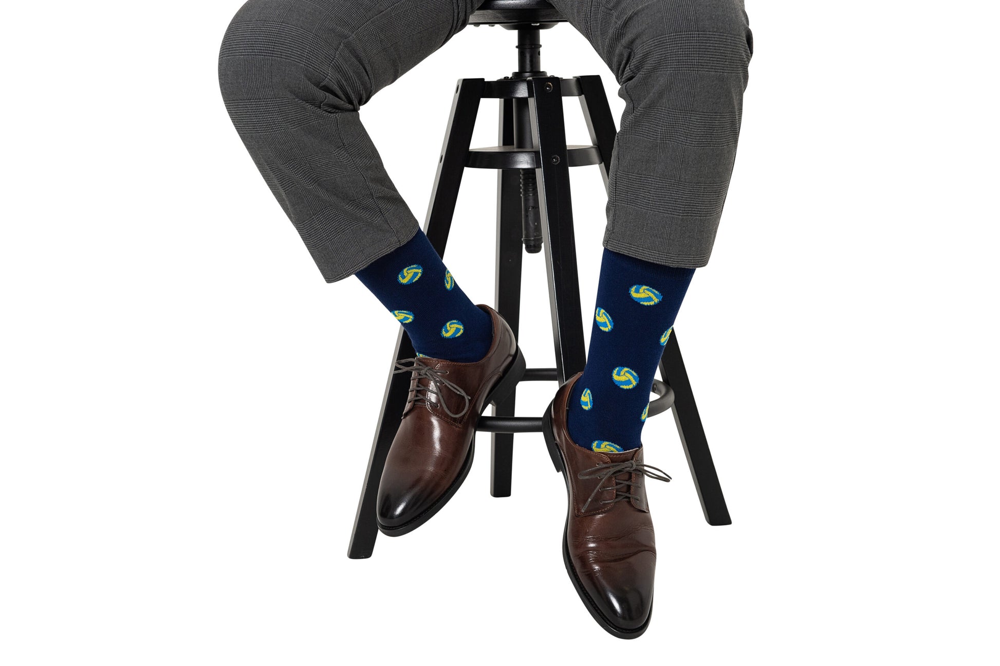 A person seated on a stool, wearing gray pants and brown dress shoes, showcases their style with Volleyball Socks in dark blue, adorned with a fashionable green and yellow spiral pattern.